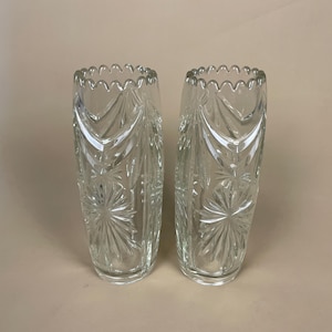May include: Two clear glass vases with a starburst and leaf design. The vases have a scalloped rim and are approximately 25 cm tall. The glass is transparent, allowing light to pass through, and the design is etched into the surface.