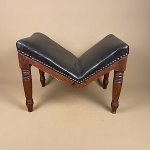 May include: A vintage V-shaped footstool featuring a black leather seat and wooden legs. The seat is edged with small metal studs. The turned legs have a dark brown finish. A classic piece of furniture, likely from a bygone era.
