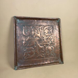 May include: Square copper tray with an embossed floral design. The tray has a hammered texture and a slightly aged patina with hints of green. The floral pattern is centered and framed within the tray's border. A decorative piece for home decor.