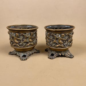 May include: Two bronze-coloured decorative planters. Each planter features a detailed relief of cherubs and foliage around the body. The planters sit on ornate, four-footed bases. The background is a solid, neutral colour.