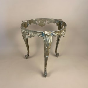 May include: An antique silver-coloured metal table base with three curved legs. The base features an ornate, openwork design around the top and along the legs. The legs are curved and end in small, rounded feet. The metal has a slightly aged appearance.