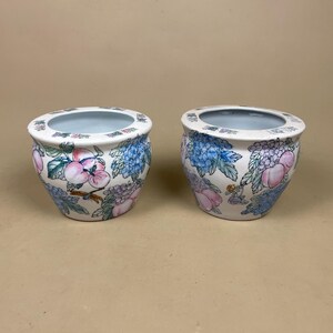 May include: Two white ceramic plant pots with floral patterns. The pots display pink, blue, and purple flowers and fruits. The rims are light blue. The pots are round with a wide opening.
