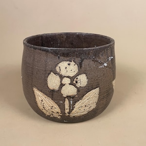 May include: A handmade, brown ceramic cup with a floral design. The cream-coloured flower has a textured appearance. The cup has a rounded shape and a dark brown rim.