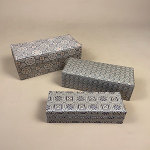 May include: Three rectangular storage boxes with lids, each featuring a distinct geometric pattern. The largest box has a light blue and white floral motif, the medium box has a dense black and white pattern, and the smallest has a blue and white grid.