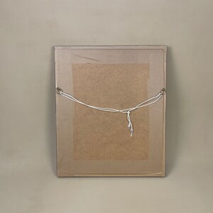 Pode incluir: A parte de tr&aacute;s de uma imagem emoldurada com um suporte de cart&atilde;o marrom e um cord&atilde;o branco preso a dois ganchos de metal.