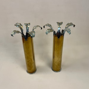 May include: Two brass vases with a floral design at the top. The vases are cylindrical with a gold finish and a silver-coloured base. The floral design has a silver-coloured finish with leaf-like details.