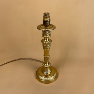 May include: A gold-toned table lamp with an ornate base and a simple lampshade holder. The lamp has a decorative design with intricate details.