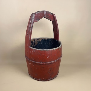 May include: An antique red wooden bucket with a curved handle. The bucket has a cylindrical shape with a dark interior and signs of wear, indicating it is a vintage item. The handle is attached to the sides of the bucket, showing signs of age.