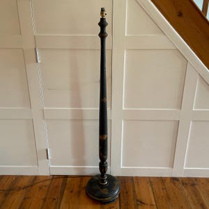 May include: A tall, slender floor lamp base featuring a matte black finish with hand-painted gold floral accents. The lamp has a turned wooden column design, a circular base, and a brass-coloured metal light socket fixture at the very top.