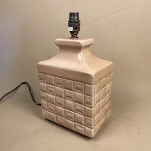 May include: A tan ceramic table lamp with a square, quilted pattern base and a rectangular top. The lamp has a brass-coloured bulb socket and a black power cord. The lamp is a neutral colour and would fit in many home decor styles.