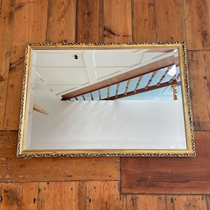 May include: A rectangular wall mirror featuring a decorative gold frame with an intricate, repeating leaf-like pattern along the border. The mirror surface is clear and reflects a view of an interior staircase with wooden railings and white walls.