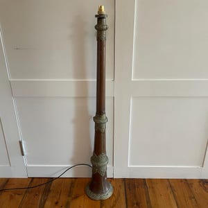 May include: A tall, decorative floor lamp featuring a dark brown wooden column and brass embellishments. The lamp has a bulb holder at the top and a flared base. It stands on a wooden floor.