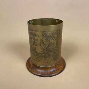May include: A cylindrical brass container with an etched Asian-inspired design, mounted on a round wooden base. The brass cup features an engraved landscape with text. The wooden base is a dark brown colour.