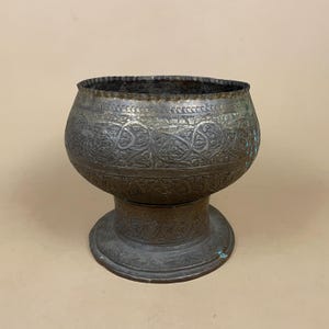 May include: A dark, weathered metal bowl with a wide, rounded body and a pedestal base. The surface features intricate, engraved patterns of swirling vines and geometric shapes, with visible signs of oxidation and aged patina throughout the metal.