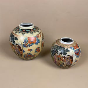 May include: Two decorative ceramic vases with a round shape. The vases feature intricate hand-painted designs with floral and figural motifs in shades of red, blue, and gold against a cream background. The vases have a black interior.