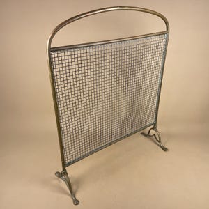 May include: An antique fireplace screen featuring a brass frame and a fine wire mesh. The screen has a curved top and stands on three decorative feet. The brass has a warm, aged patina. The mesh is a dark grey colour, adding to its vintage appeal.