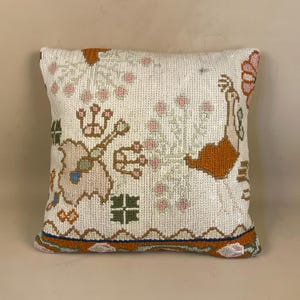 May include: Square off-white cushion with an embroidered design. The design features floral and geometric patterns in orange, pink, green, and blue. The cushion has a decorative border. The cushion measures approximately 40cm x 40cm.
