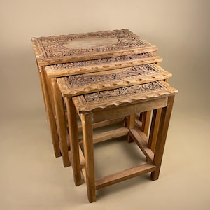 May include: A set of four nested tables crafted from light brown wood. Each rectangular table features intricate floral carvings on the top surface, a scalloped edge border, and four straight legs connected by horizontal support bars at the base.