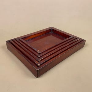 May include: A set of three rectangular nesting trays crafted from dark brown wood. Each tray features a smooth, flat base and raised edges, with the smaller trays fitting neatly inside the larger ones to create a stacked, tiered appearance.