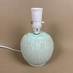 May include: A small, mint green ceramic table lamp with a textured surface. The lamp has a round base and a white lamp holder with a red switch. A white electrical cord extends from the base.