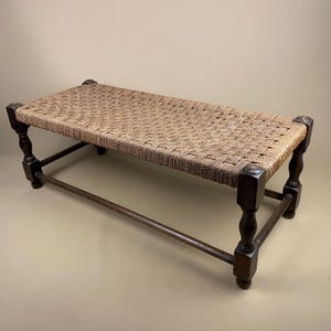 May include: A rectangular bench featuring a woven tan fibre seat supported by a dark brown wooden frame. The frame consists of four turned legs connected by horizontal stretchers, providing a stable base for the textured, woven top surface.