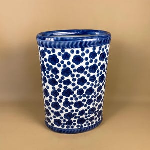 May include: A tall, cylindrical ceramic vase with a blue and white floral pattern. The vase has a dark blue rim and base, with a repeating pattern of blue flower-like shapes and dots on a white background. The vase is approximately 25 cm tall.