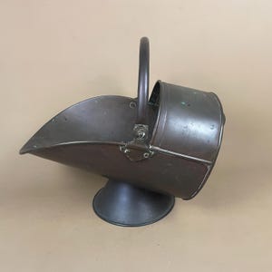May include: A dark brown metal coal scuttle featuring a wide, open-mouthed scoop design, a sturdy vertical handle secured with rivets, and a circular pedestal base. The surface displays signs of age with subtle green oxidation marks on the metal.