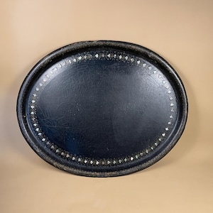 May include: Oval black serving tray with a decorative border of small, iridescent, diamond-shaped accents. The tray's surface has a cracked, aged appearance, adding to its vintage aesthetic. The rim is dark and textured, providing a contrast to the smooth interior.