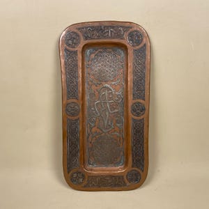 May include: A rectangular copper tray with intricate silver-toned engravings. The tray features a central design of a stylised figure with a cross, surrounded by floral motifs and geometric patterns. The edges of the tray are decorated with a repeating border of interlaced knots.