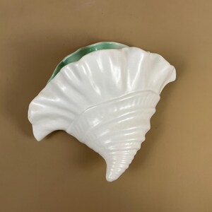 May include: A white ceramic seashell-shaped wall pocket with a glossy finish. The interior is a light green colour. The shell has detailed ridges and a spiral design, ideal for coastal decor.