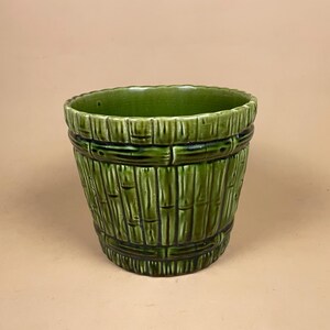 May include: A green ceramic planter with a bamboo design. The planter has a cylindrical shape with vertical bamboo stalks and horizontal bands. The glossy finish and varying shades of green create a textured appearance. Suitable for indoor or outdoor use.