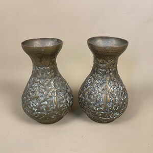 May include: Two antique brass vases with detailed floral and figure engravings. The vases have a bulbous body and a flared rim. The metal has a tarnished, aged appearance. Each vase is approximately 15 cm tall, showcasing a vintage aesthetic.
