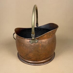 May include: A round copper coal scuttle with a dark, weathered patina and a wide, open top. It features a sturdy, arched brass handle attached with metal rivets and a small, circular brass ring handle on the side for easy lifting and transport.