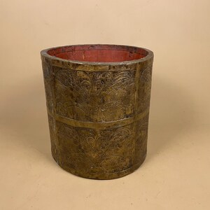 May include: A cylindrical container with a metallic gold exterior featuring embossed floral and leaf patterns arranged in a grid. The interior of the vessel is painted a solid matte red, and the structure is composed of vertical panels joined together.