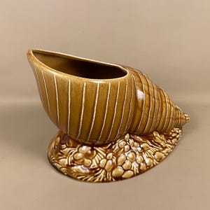 May include: A brown ceramic seashell planter with vertical white stripes. The planter has a wide opening and sits on a base with a floral design. The seashell is a decorative item.