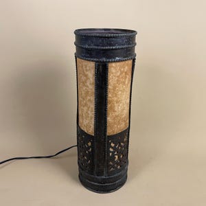 May include: A cylindrical table lamp with a black metal frame and a parchment-like shade. The lamp features decorative cutouts at the base and a textured finish. The lamp is plugged into an electrical outlet.