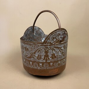 May include: A copper bucket featuring a rounded base and a high, arched handle. The exterior surface is decorated with an intricate, etched floral pattern highlighted in white, showcasing a repeating botanical motif along the upper rim area.
