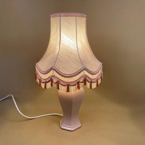 May include: A pink ceramic table lamp with a white and pink lampshade with a scalloped edge and fringe trim.