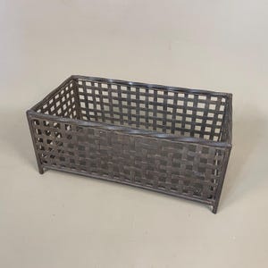 May include: A rectangular, dark grey metal basket with a woven design. The basket has a grid-like pattern on the sides and bottom, and is supported by small feet. The overall design is simple and functional.