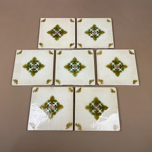 May include: A set of seven square ceramic tiles arranged in a cluster. Each tile features an off-white base with a central green and gold floral-inspired motif and small decorative corner accents. The surface of each tile has a glossy finish.