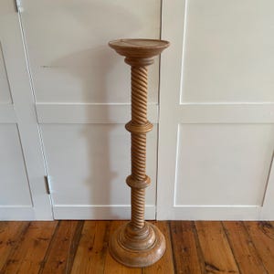 May include: A tall, light brown wooden pedestal stand featuring a circular top platform, a central column with a spiraled, twisted rope-like design, two horizontal ring accents along the shaft, and a wide, circular base for stability.