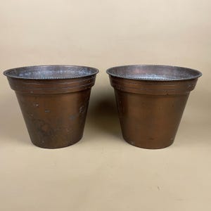 May include: Two matching tapered copper planters with a weathered patina finish. Each vessel features a wide, circular opening with a decorative beaded rim detail around the top edge, showcasing a smooth, dark metallic surface with subtle variations.