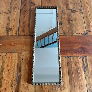 May include: A rectangular wall mirror featuring a narrow, ornate frame with a dark metallic finish and a repeating rope-like texture. The reflective glass surface shows a clear reflection of a wooden staircase railing and a white wall interior.
