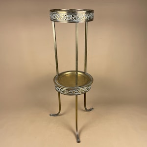 May include: A tall brass plant stand featuring two circular tiers with embossed floral patterns along the rims. The structure is supported by three slender, curved legs that extend upwards to connect the upper and lower rings, creating a vertical display.