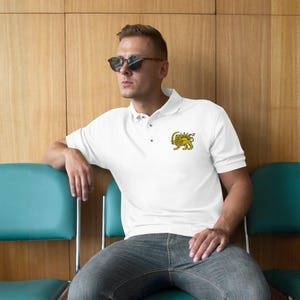 May include: White polo shirt featuring a yellow and gold lion emblem on the left breast. The shirt has a classic collar and short sleeves. The person is wearing sunglasses and sitting on a teal chair.