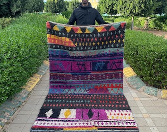 May include: A colorful Moroccan rug with geometric patterns in shades of red, blue, yellow, and purple. The rug has a fringe border and is being held up by a person.