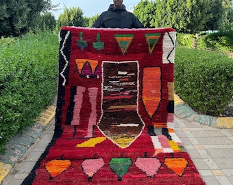 May include: A red Moroccan rug with a geometric pattern featuring abstract shapes, hearts, and a central design with a brown and white striped pattern. The rug has a white fringe border.