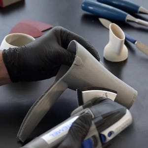 Puede incluir: Una persona con guantes negros lija un objeto de cer&aacute;mica gris con una herramienta el&eacute;ctrica. La herramienta tiene un mango negro y azul y un disco de lijado gris. El objeto tiene forma de embudo o cono.