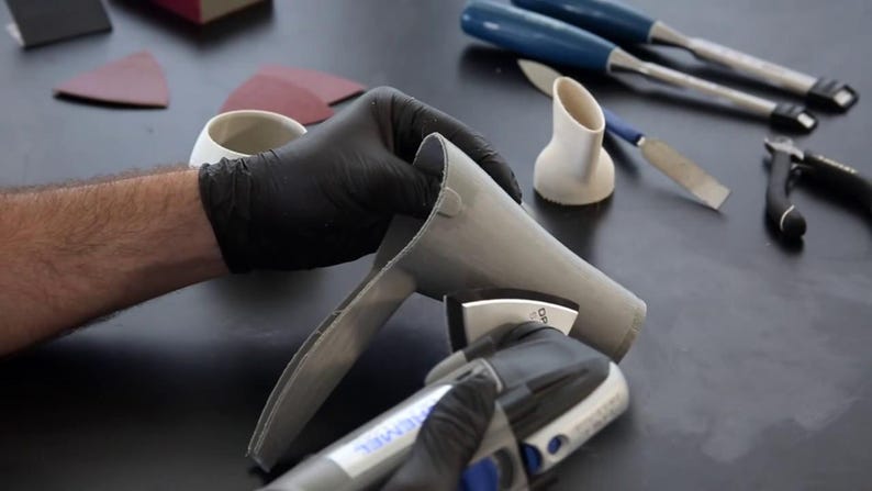Puede incluir: Una persona con guantes negros lija un objeto de cer&aacute;mica gris con una herramienta el&eacute;ctrica. La herramienta tiene un mango negro y azul y un disco de lijado gris. El objeto tiene forma de embudo o cono.