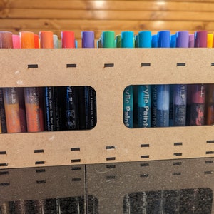 Könnte beinhalten: Eine Holzkiste mit zwei durchsichtigen Fenstern, die Reihen von bunten Acryl-Farbstiften zeigen. Die Stifte sind mit "Acrylic Paint" und "MILE" beschriftet.